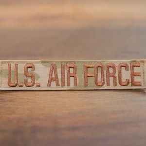 May include: A tan and green camouflage fabric patch with the words "U.S. AIR FORCE" embroidered in copper-colored thread.