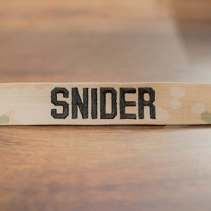 May include: A beige camouflage fabric name tag with the name "SNIDER" embroidered in black.