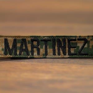 Könnte beinhalten: Ein rechteckiger Namensaufnäher mit dem Namen "MARTINEZ" in schwarzer Schrift. Der Aufnäher hat ein Tarnmuster in Grün-, Braun- und Beigetönen. Er ist wahrscheinlich für militärische oder taktische Zwecke bestimmt.