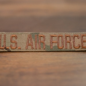May include: A brown and green camouflage fabric patch with the text "U.S. AIR FORCE" embroidered in brown thread.
