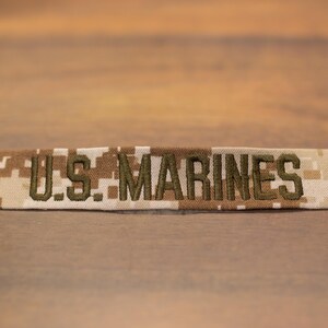 Könnte beinhalten: Ein rechteckiger Aufnäher mit dem Text "U.S. MARINES" in olivgrünem Faden gestickt. Der Aufnäher hat ein Tarnmuster in Beige-, Braun- und Weißtönen. Der Aufnäher ist wahrscheinlich für Militäruniformen.