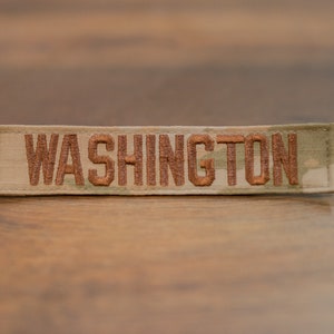 Può includere: Un patch in tessuto mimetico beige e verde con la parola "WASHINGTON" ricamata in filo marrone.