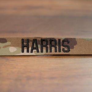 Könnte beinhalten: Ein Namensschild aus Tarnstoff mit dem Namen "HARRIS", der in schwarzem Garn gestickt ist.