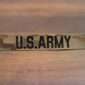 May include: A camouflage fabric patch with the text "U.S. ARMY" embroidered in black.