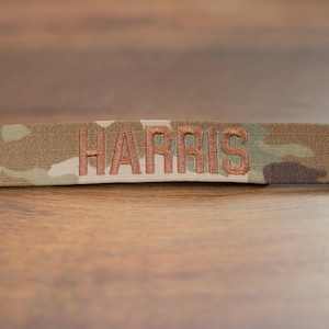 以下が含まれることがあります： 茶色、緑、ベージュの色合いのカモフラージュ柄の長方形パッチ。中央には「HARRIS」の文字が銅色の糸で刺繍されています。このパッチは、軍事用または戦術的な装備品に使用される可能性があります。サイズはセンチメートルです。