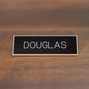May include: A black rectangular name tag with white text that reads "DOUGLAS".