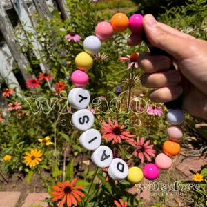 May include: A colorful beaded bracelet with the word "DAISY" spelled out in white beads. The bracelet is made with pink, yellow, orange, and white beads.