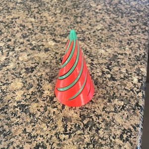 May include: A red and green decorative cone-shaped object. The cone has a green tip and green curved lines that wrap around the red body. The object is likely a Christmas decoration or part of a festive display.