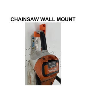 Op de afbeelding: Een oranje kettingzaag muurbevestiging met een oranje en grijze kettingzaag. De kettingzaag heeft een zwarte handgreep en de tekst "STIHL" is zichtbaar. De tekst "CHAINSAW WALL MOUNT" staat boven de afbeelding.