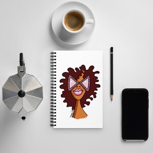 May include: A spiral bound notebook with a white cover featuring a cartoon illustration of a woman with brown hair wearing pink and green patterned sunglasses. The woman is smiling.