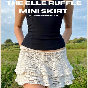 May include: A cream-colored, crochet mini skirt with a ruffled hem. The skirt is paired with a black halter top. Text on the image reads "THE ELLE RUFFLE MINI SKIRT" and "PATTERN BY @HOOKEDBYELLE". The outfit is worn outdoors.