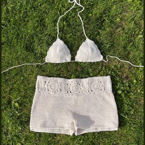Może przedstawiać: Biały zestaw plażowy z szydełka, składający się z trójkątnego topu od bikini i szortów z wysokim stanem. Szorty mają dekoracyjny kwiatowy wzór na pasie. Tekst "The WILLOW beach set" jest widoczny.