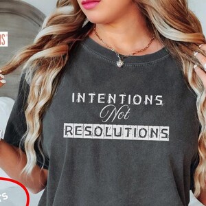 May include: A dark grey t-shirt with the text "INTENTIONS Not RESOLUTIONS" printed in white. The shirt is being worn by a person sitting on a bed. The text "COMFORT COLORS" is visible in the top left corner of the image.