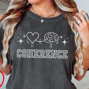 May include: A dark gray t-shirt with a white graphic of a heart and brain holding hands. The text "COHERENCE" is printed below the graphic. The shirt is being modeled by a person.