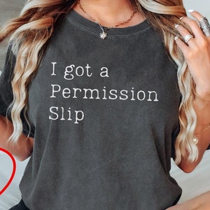 Könnte beinhalten: Ein graues T-Shirt mit dem Text "I got a Permission Slip" auf der Vorderseite. Das Shirt wird von einer Person getragen, die auf einem Bett sitzt. Der Text "More Colors available" ist in einem roten Kreis auf dem Bett geschrieben.
