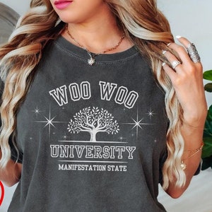 Könnte beinhalten: Ein dunkelgraues T-Shirt mit einem weißen Grafikdesign, auf dem "Woo Woo University Manifestation State" steht. Die Grafik zeigt einen stilisierten Baum mit Sternen auf beiden Seiten.