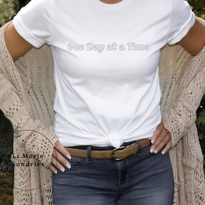 Peut inclure: T-shirt blanc avec le texte "One Day at a Time" imprimé sur le devant. Le t-shirt est porté par une personne avec un cardigan beige et un jean bleu.