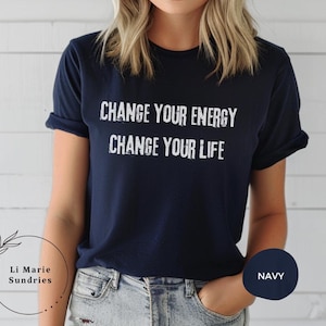 May include: Navy blue t-shirt with the text "CHANGE YOUR ENERGY CHANGE YOUR LIFE" in white. The shirt is a crew neck and has rolled-up sleeves. The shirt is a solid color and is being worn.