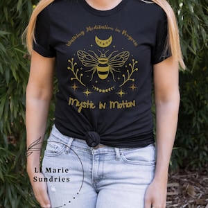 Può includere: T-shirt nero con grafica dorata di un'ape, luna crescente e stelle, con le scritte "Walking Meditation in Progress" e "Mystic in Motion". La maglietta è annodata in vita.