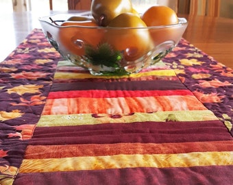 May include: A decorative table runner with a patchwork design featuring various colors, including burgundy, orange, and green. A clear glass bowl filled with oranges sits on top of the runner. The runner is approximately 12 inches wide.