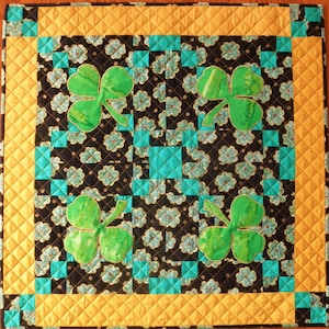 May include: A black and gold fabric quilt with a green shamrock design. The quilt is quilted in a diamond pattern and has a gold border.