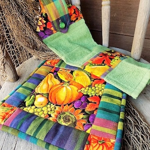 May include: A green kitchen towel with a colorful autumn-themed fabric panel attached. The panel features pumpkins, grapes, and sunflowers. The towel is hanging on a wooden chair with a woven net behind it.