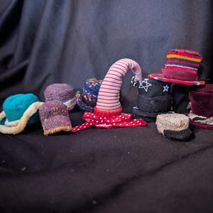 Könnte beinhalten: Eine Sammlung von Miniaturhüten in verschiedenen Stilen und Farben. Enthält einen türkisfarbenen Hut mit cremefarbenem Besatz, einen rot-weiß gestreiften Hut und einen schwarzen Zylinder mit Sternmuster. Weitere Hüte sind bordeauxrot, grau und mehrfarbig.