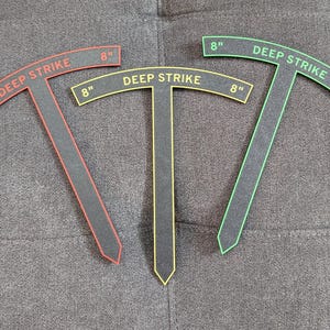 Op de afbeelding: Drie T-vormige spelsjablonen van zwart materiaal met gekleurde omlijningen en tekst. Elk sjabloon is voorzien van de tekst DEEP STRIKE en 8" op het oppervlak. De omlijningen en de tekst zijn respectievelijk in rood, geel en groen uitgevoerd.