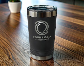 Pode incluir: Um copo de aço inoxidável preto fosco com borda prateada e tampa transparente. A frente apresenta um design de logotipo circular gravado em prata acima do texto "YOUR LOGO ETCHED HERE" em uma fonte sem serifa, com um design elegante.