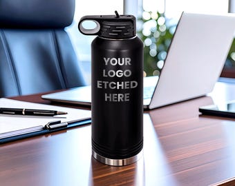 Peut inclure: Une bouteille d'eau cylindrique noir mat avec un couvercle noir doté d'une boucle de transport. L'avant de la bouteille porte le texte "YOUR LOGO ETCHED HERE" en gris clair. La base de la bouteille présente une fine bande métallique argentée.