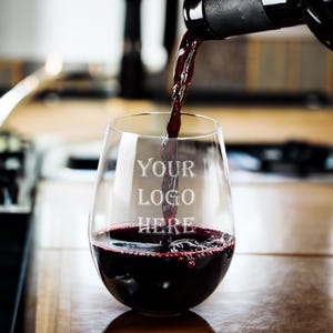 May include: A stemless wine glass being filled with red wine from a dark bottle. The glass has the text "YOUR LOGO HERE" etched on the front. The wine is a deep red color, and the glass is sitting on a wooden surface.