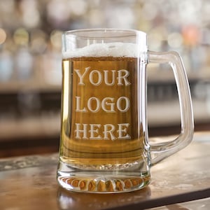 Könnte beinhalten: Ein klarer Bierkrug aus Glas mit einem stabilen Henkel, gefüllt mit einer goldgelben Flüssigkeit und einer weißen Schaumkrone. Auf der Vorderseite des Glases befindet sich der gravierte Text "YOUR LOGO HERE" in einer Serifenschrift.