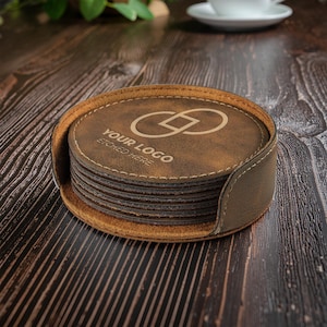 May include: A set of brown leather coasters with a holder. The oval coasters have a stitched border and the text "YOUR LOGO ETCHED HERE" and a logo design. The holder is a darker brown colour.