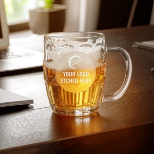 Puede incluir: Una jarra de cerveza de vidrio transparente llena de cerveza y espuma. La jarra tiene un asa y un diseño grabado en la parte delantera que dice "YOUR LOGO ETCHED HERE". La jarra está sobre una superficie de madera oscura.