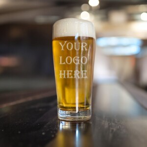 May include: A clear glass pint beer glass filled with golden lager and a thick white foam head. The glass features the etched white text YOUR LOGO HERE centered on the front. The vessel has a tapered cylindrical shape with a thick base.