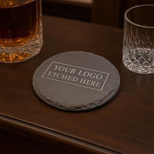 May include: A round, dark gray slate coaster with the text "YOUR LOGO ETCHED HERE" in a white rectangular frame. The coaster sits on a dark wooden surface, with a glass of amber liquid and a clear glass in the background.