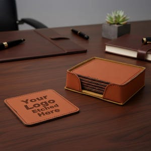 May include: A set of square, brown leather coasters with the text "Your Logo Etched Here" etched on one. The coasters are in a matching holder. The set is on a wooden desk with other leather office accessories.