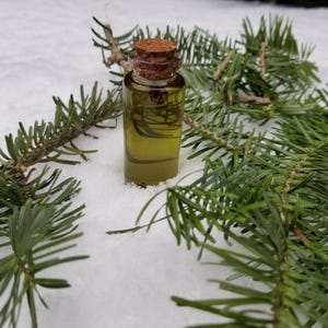 May include: A small, clear glass bottle with a cork stopper, filled with a yellow-green liquid, rests on snow. Green pine branches surround the bottle, creating a natural, wintery scene. The bottle appears to contain essential oil.