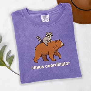 May include: A purple short-sleeve t-shirt featuring a graphic of a raccoon riding on the back of a brown bear. Below the bear, the text reads "chaos coordinator" in light yellow letters. The shirt has a Comfort Colors brand neck label.