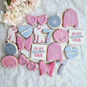 May include: A selection of decorated sugar cookies in various shapes, including heart-shaped sunglasses, butterflies, disco balls, and jackets. The cookies are primarily pink and blue, with the text "IN MY BIRTHDAY ERA" and names on some. The cookies are arranged on a white surface.