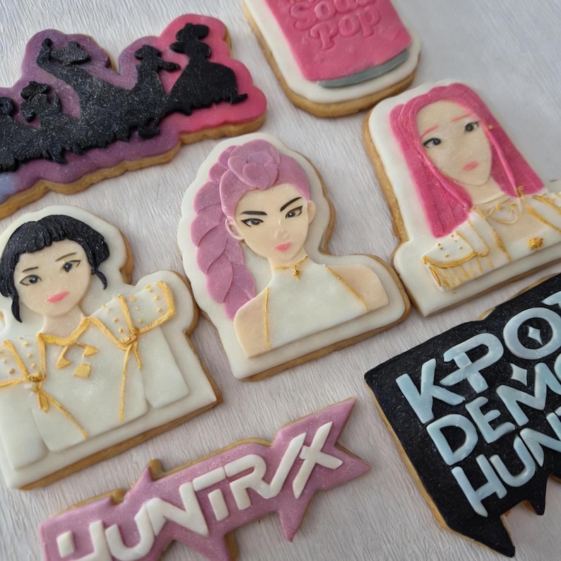 Puede incluir: Surtido de galletas de az&uacute;car decoradas con dise&ntilde;os tem&aacute;ticos de K-pop. Las galletas incluyen retratos de mujeres con cabello rosado, una lata de refresco rosa y los textos "KPOP DEMON HUNTERS" y "HUNTR/X".