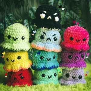 Può includere: Una pila di nove peluche all'uncinetto in vari colori, tra cui verde, giallo, blu, rosso, viola e blu chiaro. Ogni peluche ha un'espressione facciale diversa e presenta un design all'uncinetto.