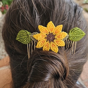 May include: A handmade hair comb featuring a yellow and brown sunflower design, with green leaves on either side. The sunflower is the focal point, with detailed petals and a dark brown center. The comb has gold-colored metal prongs.