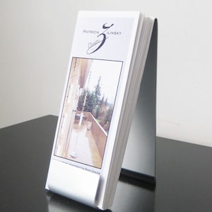 May include: A white brochure featuring the text "Patricia Ilinsky Designs" and a photograph of a patio with a rain chain. The brochure is presented in a silver stand on a black surface.