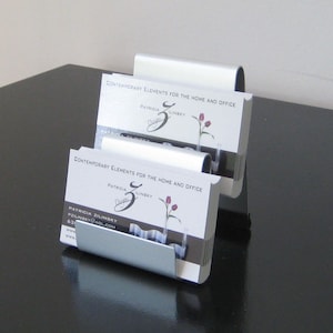 May include: Two silver business card holders with a stack of business cards. The business cards are white with black text and a pink flower design. The text on the business cards reads "CONTEMPORARY ELEMENTS FOR THE HOME AND OFFICE", "PATRICIA ZILINSKY", and "Design".