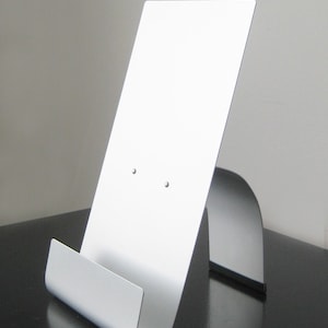 May include: A silver metal book stand with a curved base and a flat top. The stand is designed to hold a book or tablet upright.