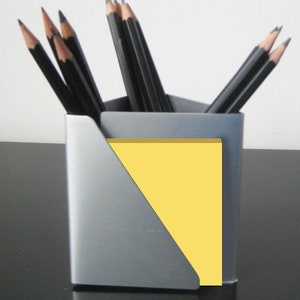 May include: A silver pencil holder with a yellow sticky note pad. The holder has a triangular opening for the note pad. There are multiple black pencils in the holder.