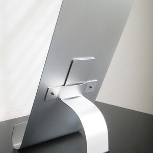 May include: A silver metal picture stand with a curved base and a metal bracket to hold the picture in place.