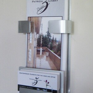 May include: A silver wall mounted brochure holder with two brochures. The top brochure has the text "Patricia Ilinsky Designs" and a stylized letter "Z". The bottom brochure has the text "Contemporary Elements for the Home and Office" and a stylized letter "Z".