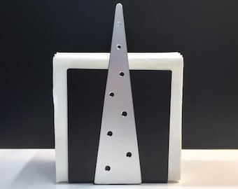 May include: A silver metal napkin holder with a triangular design and circular cutouts. The holder is positioned in front of a stack of white napkins and a black base, creating a modern and minimalist aesthetic. The design is sleek and suitable for various decor styles.